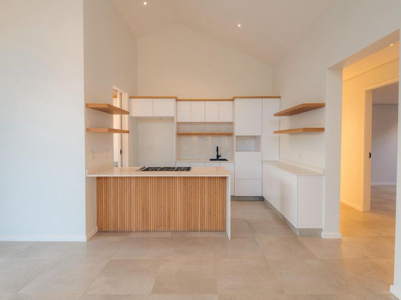Modern kitchen featuring white cabinetry, timber slat island, stone countertops, gas hob, and floating shelves under a high vaulted ceiling.