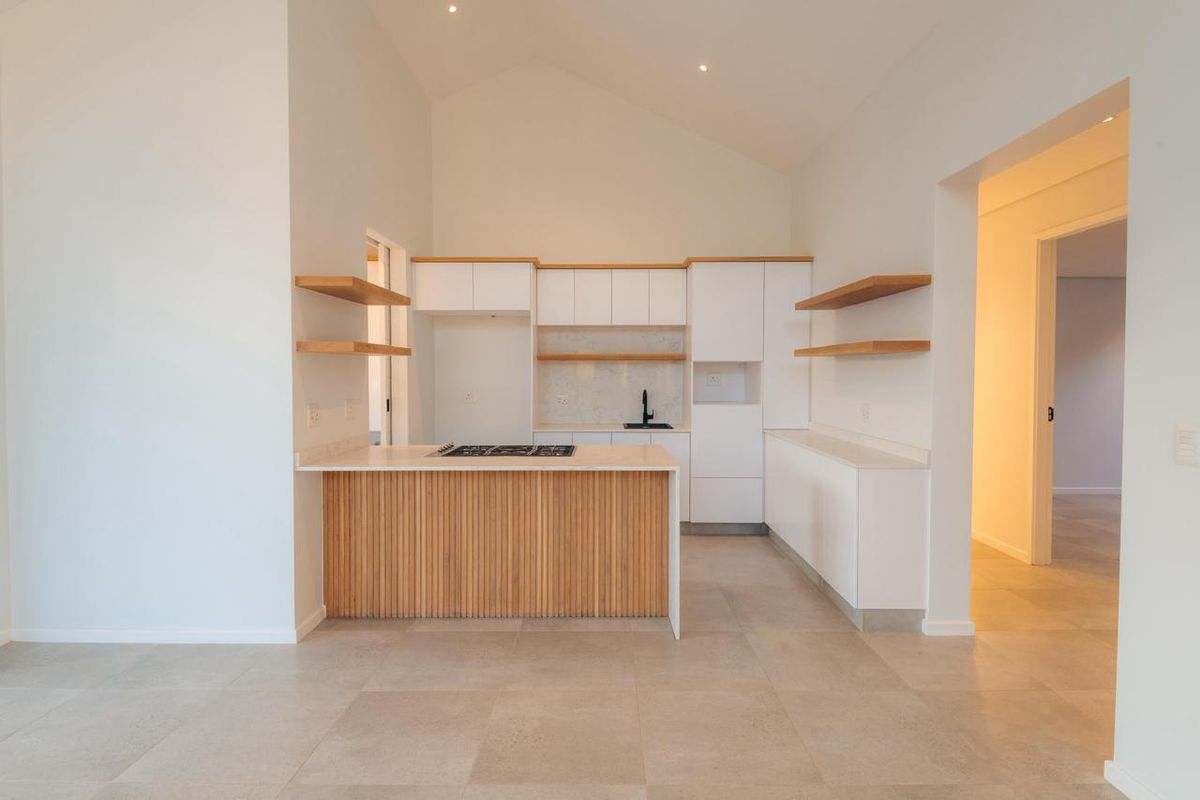 Modern kitchen featuring white cabinetry, timber slat island, stone countertops, gas hob, and floating shelves under a high vaulted ceiling.