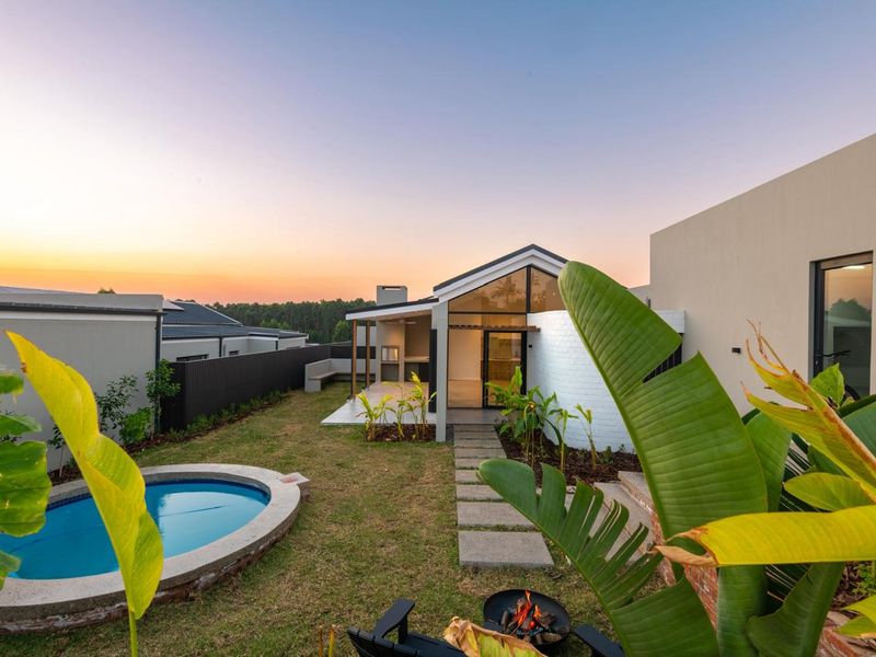 A modern rear exterior view featuring a landscaped garden, a small swimming pool, a covered patio with a built-in braai, and a contemporary glass facade.