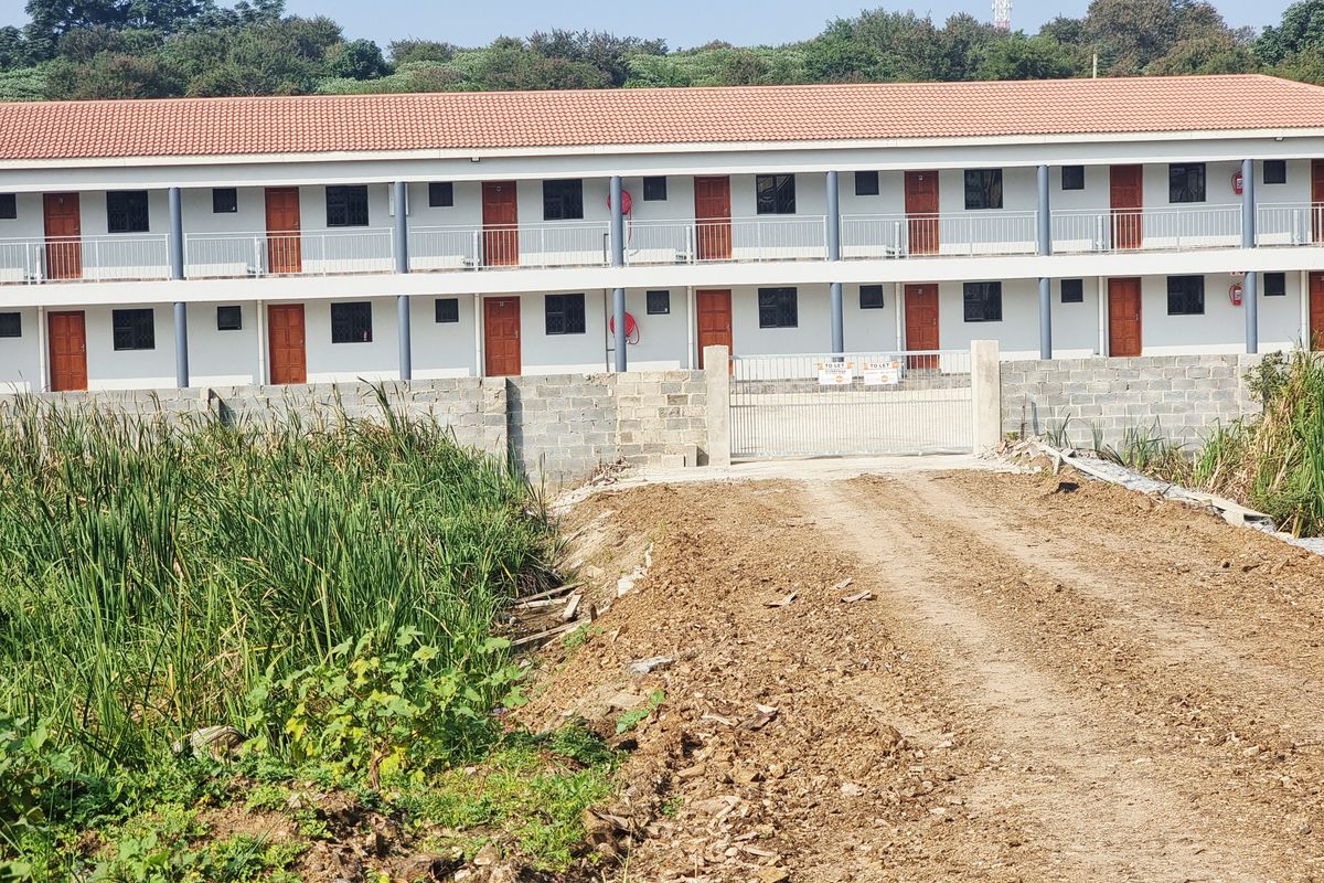 Exterior view of a multi-level residential complex featuring a tiled roof, balcony access, and a gated entrance with an unpaved approach.