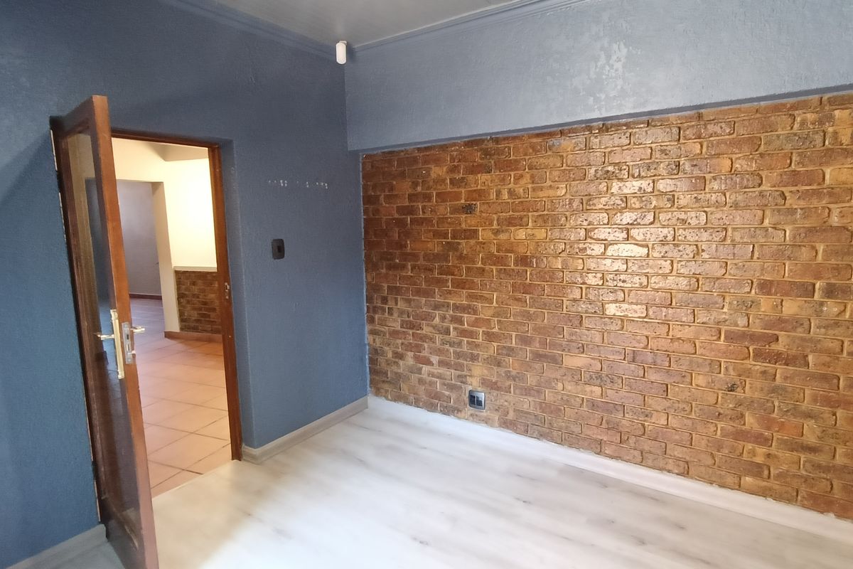 A bedroom featuring a prominent exposed brick feature wall, light-toned laminate flooring, and a wooden door leading to an adjacent living area.
