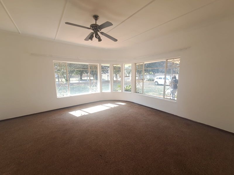 A spacious, empty living room featuring a large bay window with security bars, a ceiling fan, and neutral carpeting.