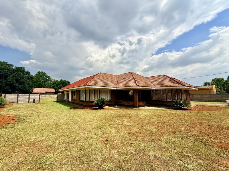 A rear view of a single-story brick house featuring a terracotta tiled roof, large windows, and a surrounding garden with a concrete patio area.