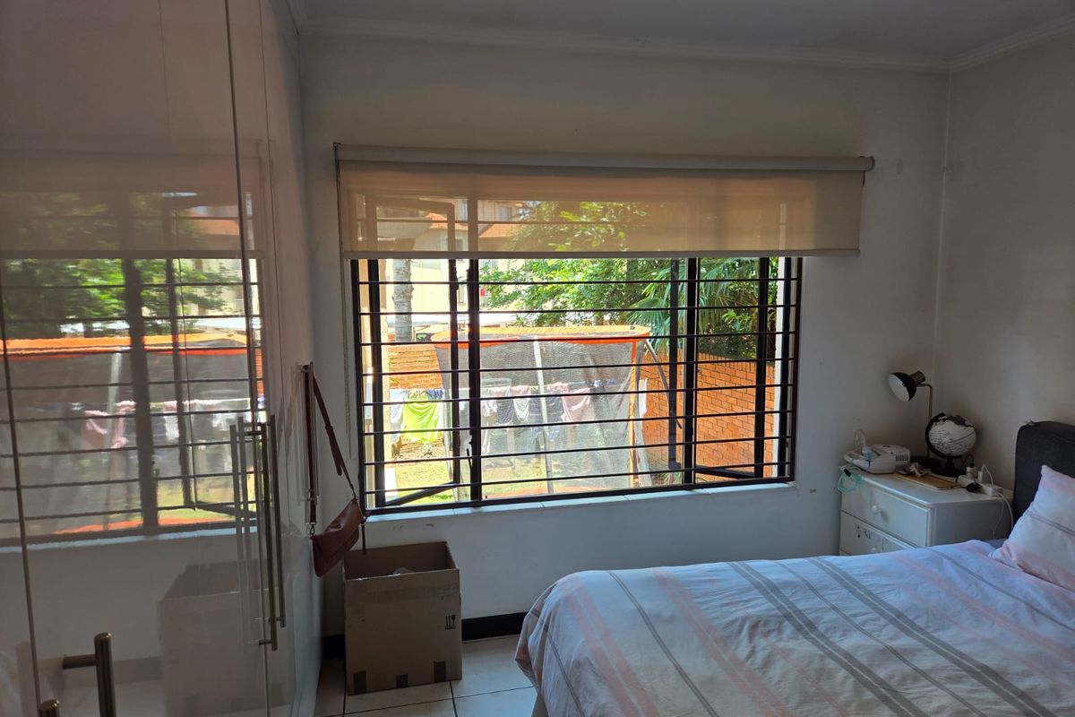 A bedroom featuring built-in cupboards, tiled flooring, and a large window with security bars overlooking the rear garden area.