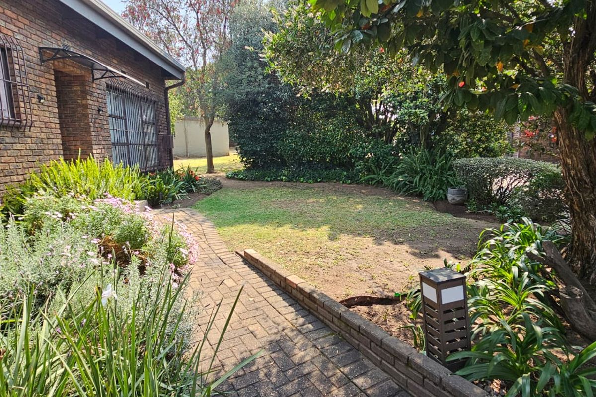 A view of the townhouse exterior featuring a brick facade, paved walkway, and established garden area with mature trees and shrubs.