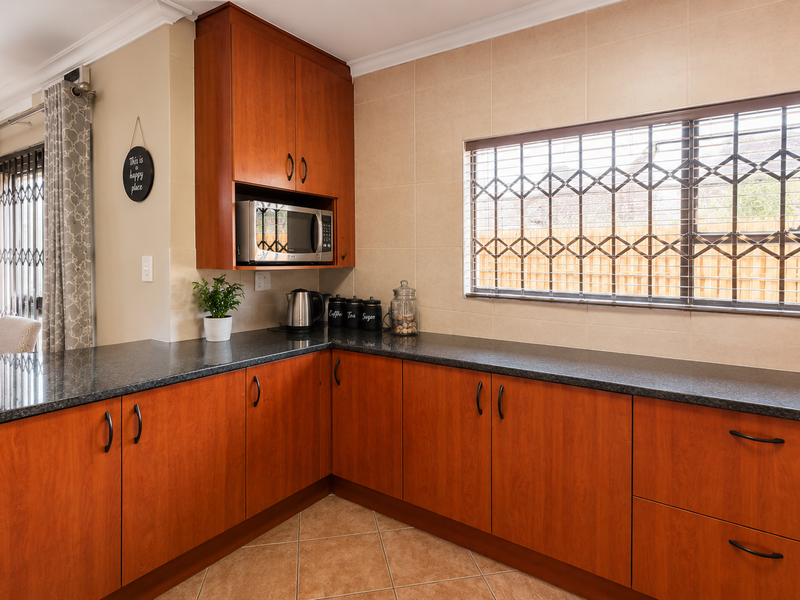 A functional kitchen featuring warm-toned wooden cabinetry, dark granite countertops, a tiled backsplash, and a window fitted with a security gate.