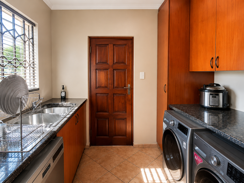 A functional laundry room featuring granite countertops, double stainless steel sink, ample wooden cabinetry, and tiled flooring with security bars on the window.