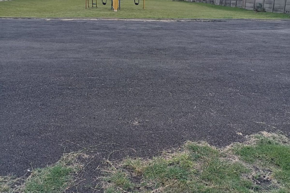 A communal outdoor area featuring a paved asphalt section and a grass lawn with a swing set, enclosed by perimeter boundary walls.