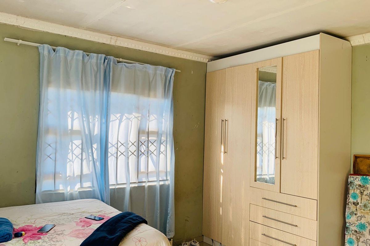 A bedroom featuring a built-in wardrobe, security bars on the window, and decorative ceiling cornice. The walls show signs of wear and paint damage.