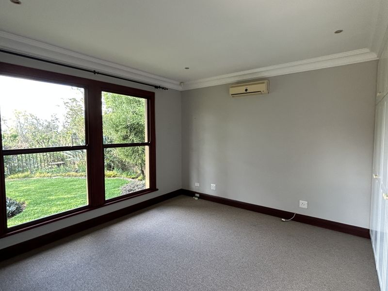 A bright, empty bedroom featuring neutral walls, carpeted flooring, built-in cupboards, an air conditioning unit, and large windows overlooking the garden.
