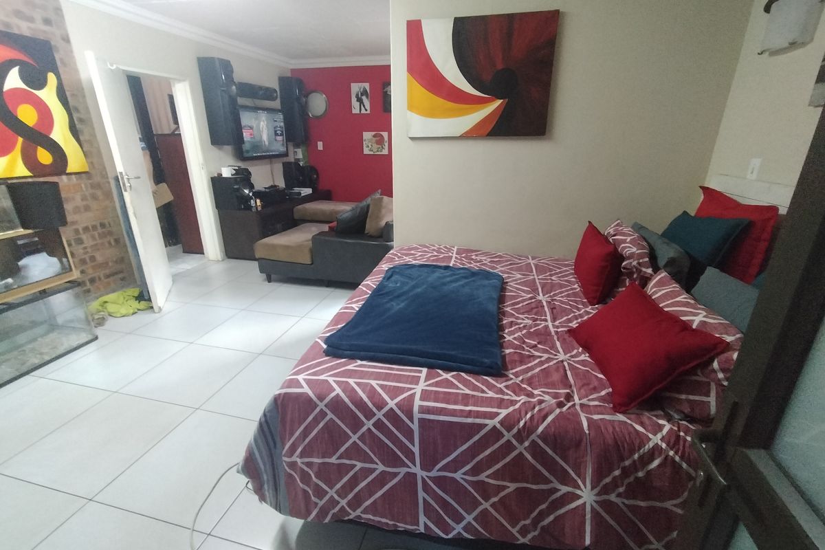 A bedroom featuring tiled flooring and an exposed brick feature wall, with an open layout connecting to an adjacent living area.