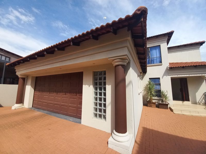 Front elevation of a residential property featuring a double garage, decorative columns, terracotta roof tiles, and a paved driveway entrance.