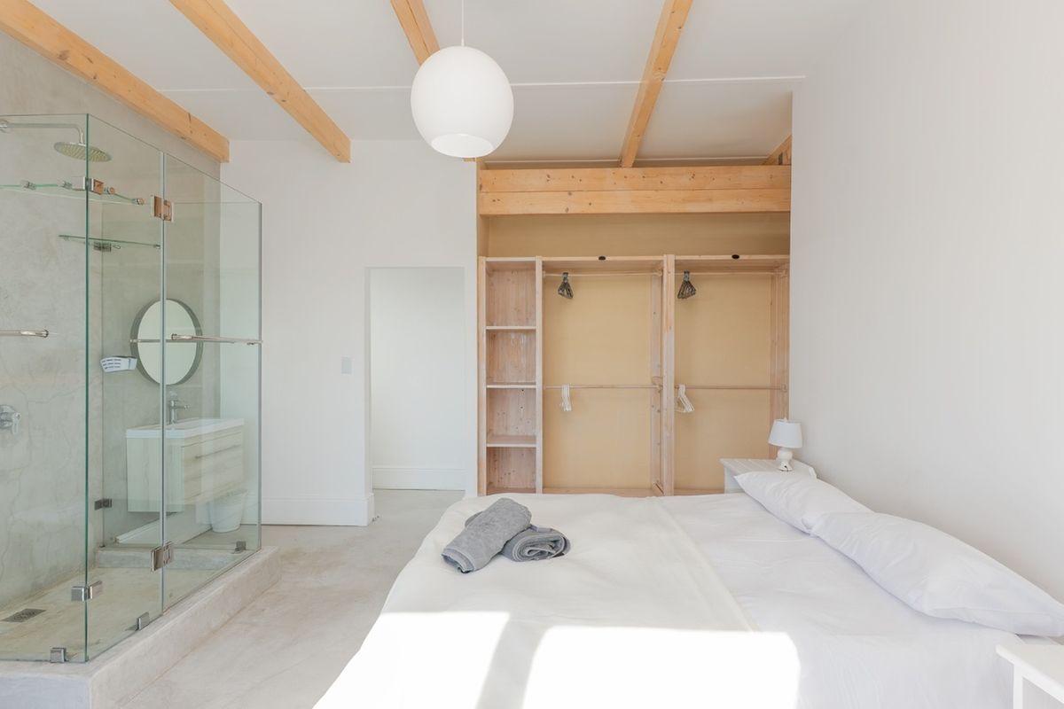 Modern bedroom featuring exposed timber ceiling beams, polished concrete floors, and an integrated glass-enclosed shower and vanity area with built-in wardrobe storage.