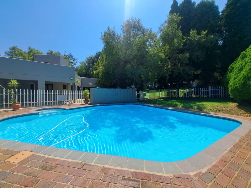 A well-maintained outdoor swimming pool area featuring brick coping, a white picket fence, and surrounding landscaped garden space.