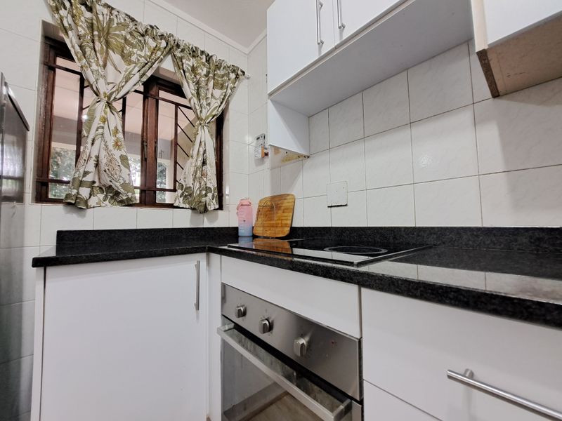 Compact kitchen featuring white cabinetry, dark granite countertops, built-in oven, and electric hob with tiled backsplash and window security bars.