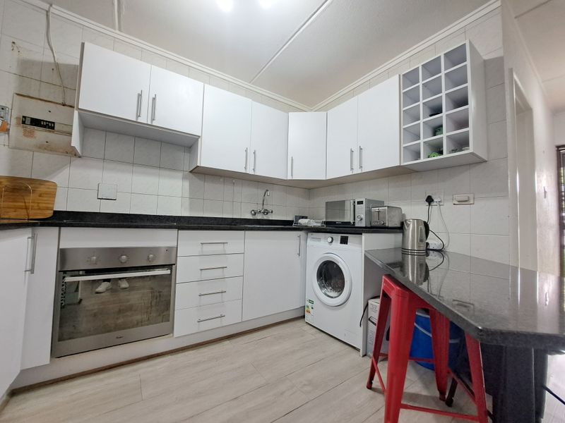 Compact kitchen featuring white cabinetry, granite countertops, built-in oven, and an integrated washing machine with tiled backsplash and wood-look flooring.