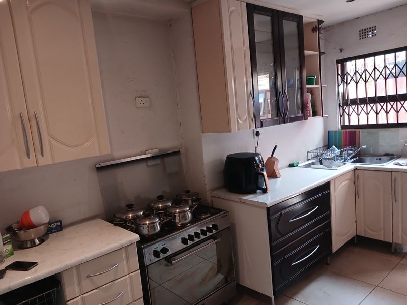 A functional kitchen featuring built-in cabinetry, a freestanding stove, and a stainless steel sink, with visible wear on wall finishes and cabinetry.