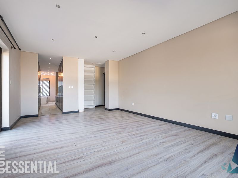 A spacious, empty living area featuring modern laminate flooring, recessed downlighting, and neutral wall tones, leading into an adjacent hallway space.