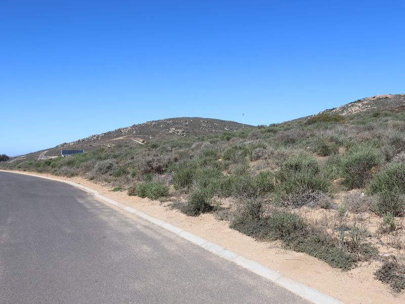 A view of the asphalt road and concrete curbing bordering the vacant residential land with natural hillside terrain.