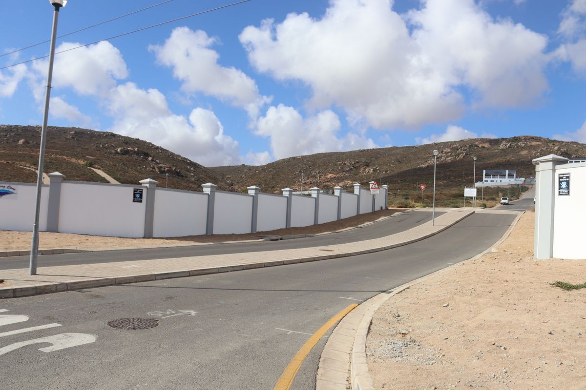 A view of the paved street and sidewalk bordering a residential property with a white perimeter wall and surrounding hilly landscape.