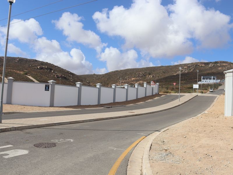 A view of the paved street and sidewalk bordering a residential property with a white perimeter wall and surrounding hilly landscape.