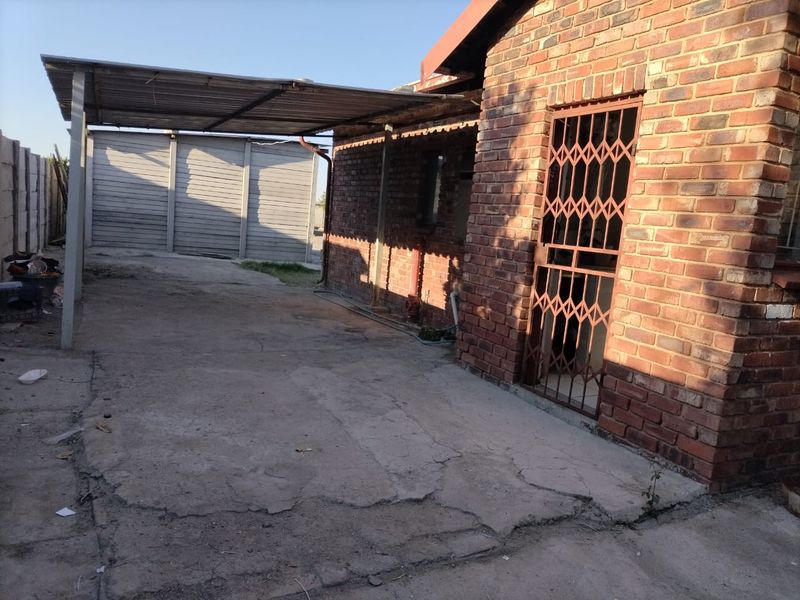 Rear exterior view of the property featuring a face brick facade, a metal carport structure, and a concrete driveway with visible surface cracking.