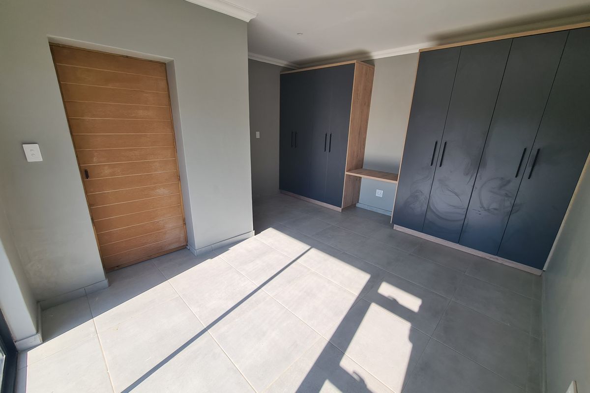 A clean, empty bedroom featuring modern grey tiled flooring, built-in dark cabinetry with integrated desk space, and a solid timber door.