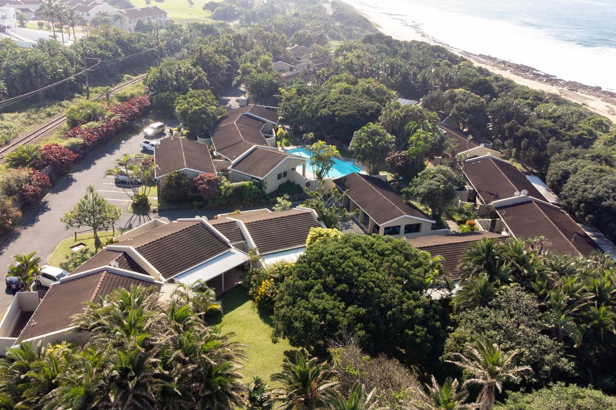 Aerial view of a coastal townhouse complex featuring tiled roofs, landscaped gardens, a communal swimming pool, and proximity to the beach.