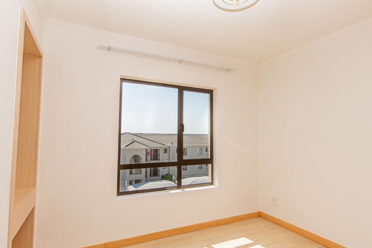 A bright, empty bedroom featuring light-toned tiled flooring, a built-in wooden shelving unit, and a window with a view of the surrounding complex.