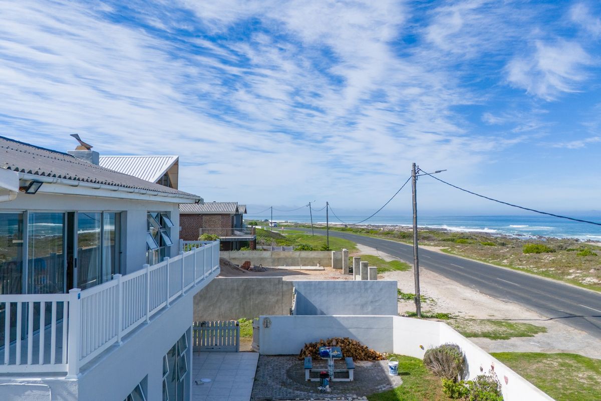 Elevated view of the rear exterior featuring a balcony, paved patio area, and boundary wall, overlooking the coastal road and ocean.