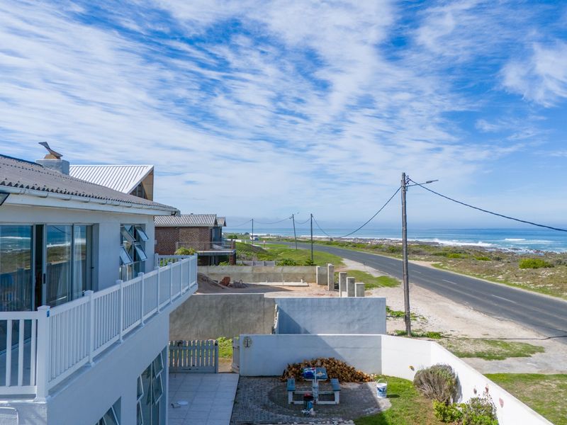 Elevated view of the rear exterior featuring a balcony, paved patio area, and boundary wall, overlooking the coastal road and ocean.