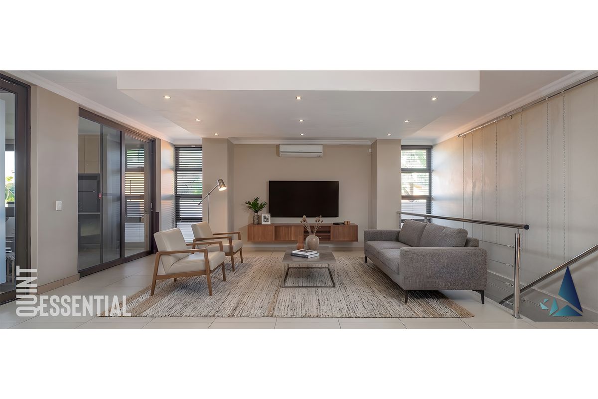 A modern, well-lit living room featuring tiled floors, recessed ceiling lighting, a split-system air conditioner, and a sleek stainless steel staircase railing.