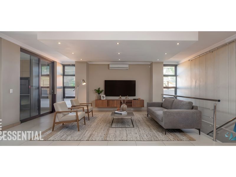 A modern, well-lit living room featuring tiled floors, recessed ceiling lighting, a split-system air conditioner, and a sleek stainless steel staircase railing.