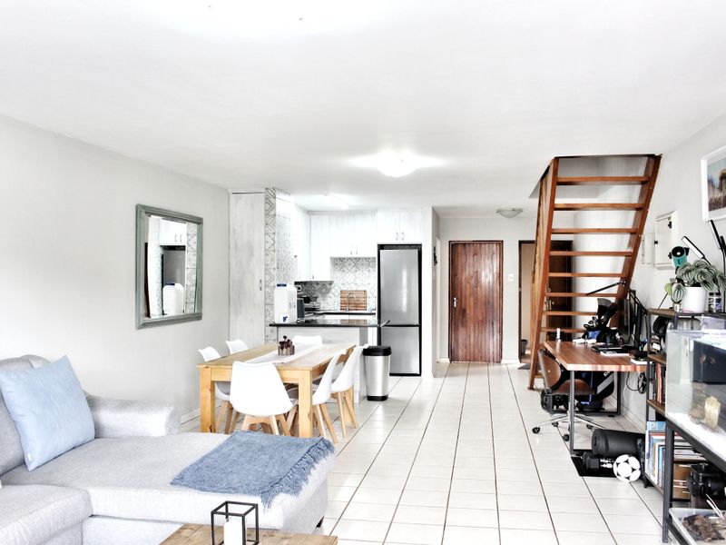 Open-plan living area featuring tiled flooring, a wooden staircase, and a functional kitchen with cabinetry and a tiled backsplash.