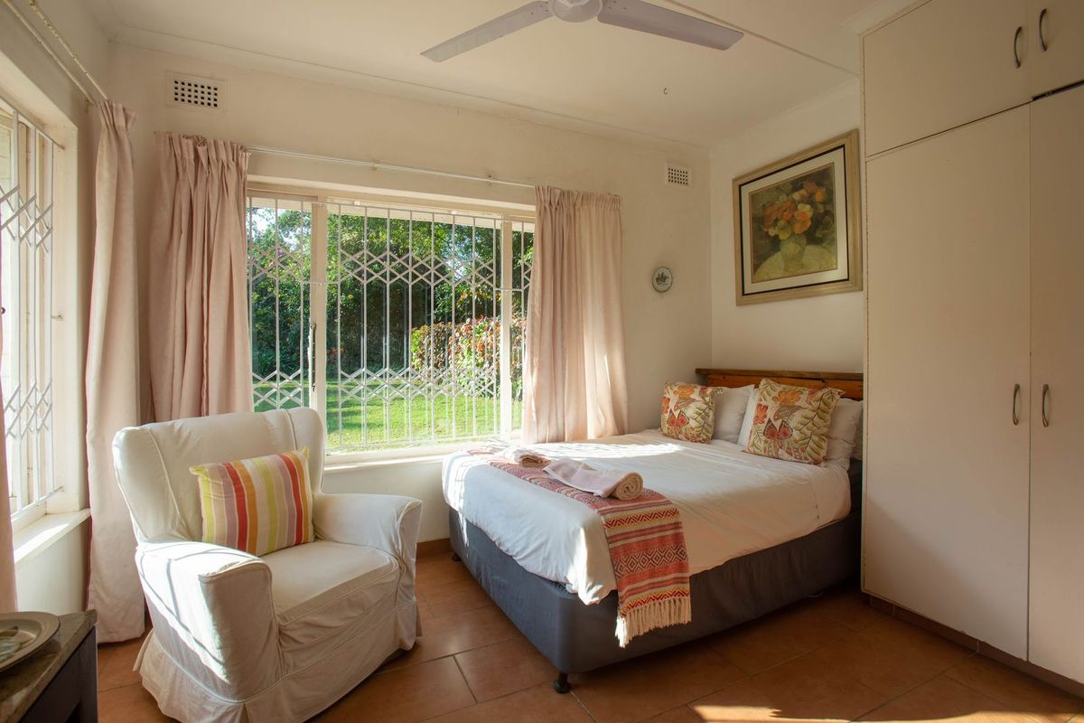 A bedroom featuring tiled flooring, built-in cupboards, a ceiling fan, and large windows with security bars overlooking the garden.