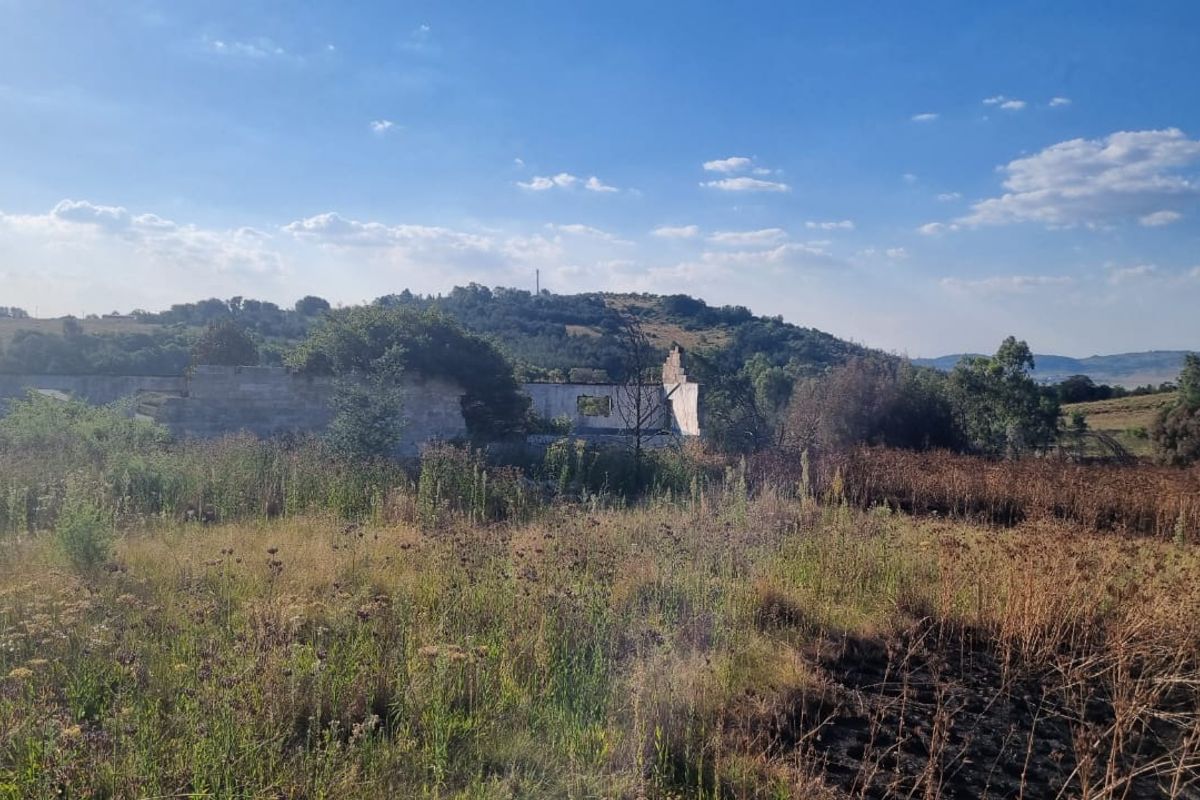 A view of a vacant residential plot featuring the remains of a ruined structure amidst overgrown vegetation and natural landscape.