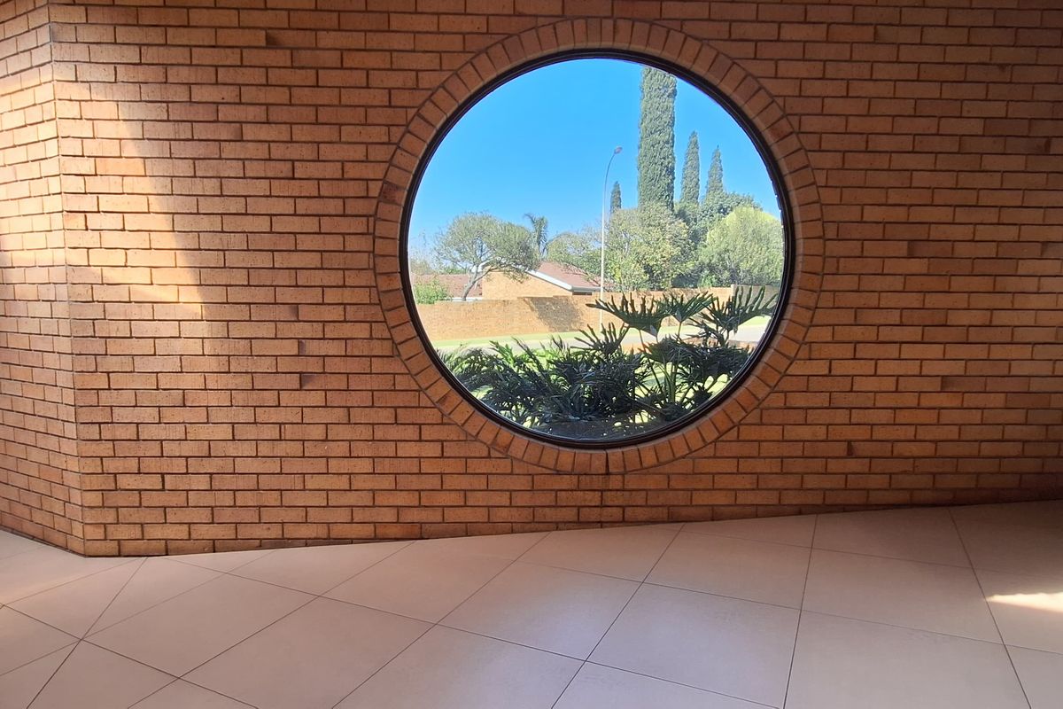 Spacious living area featuring a distinctive circular window, exposed brick walls, and neutral tiled flooring, offering a bright and unique architectural aesthetic.