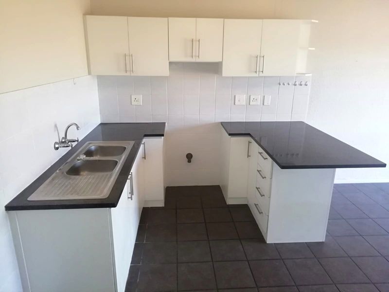 A clean, empty kitchen featuring white built-in cabinetry, dark granite countertops, a double stainless steel sink, and tiled flooring.