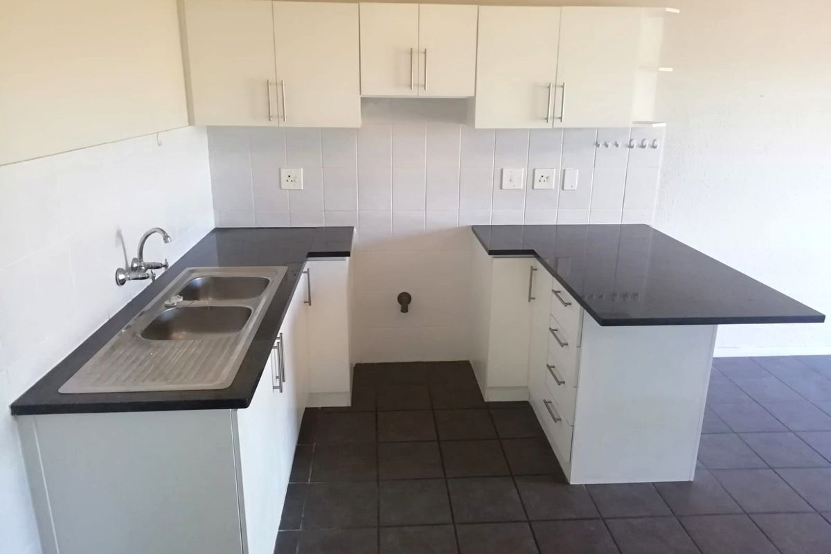 A clean, empty kitchen featuring white built-in cabinetry, dark granite countertops, a double stainless steel sink, and tiled flooring.