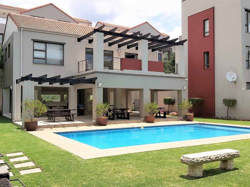 A well-maintained communal pool area featuring a covered patio with pergola, paved surround, and landscaped garden within an apartment complex.