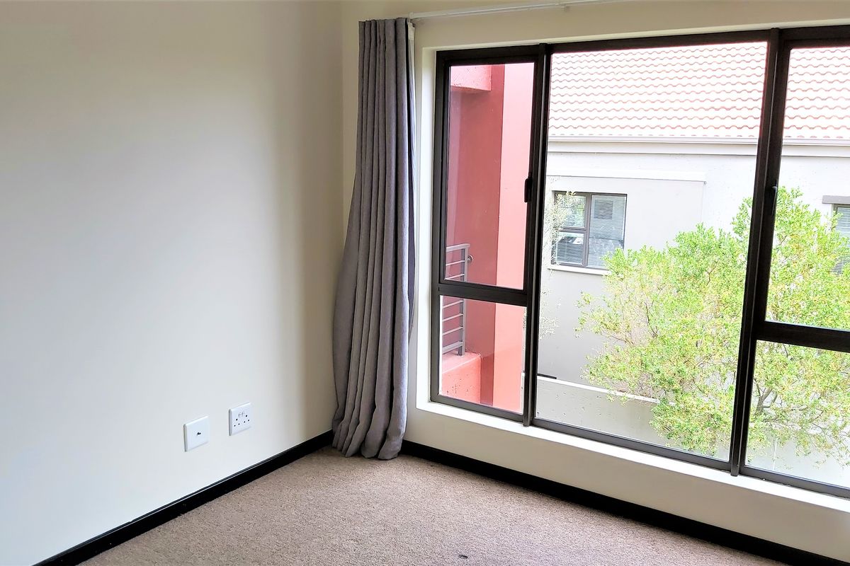 A bright, empty bedroom featuring neutral walls, carpeted flooring, and large aluminium-framed windows with views of the surrounding complex.