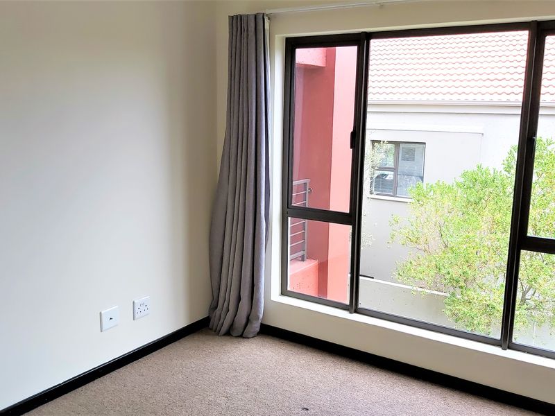 A bright, empty bedroom featuring neutral walls, carpeted flooring, and large aluminium-framed windows with views of the surrounding complex.