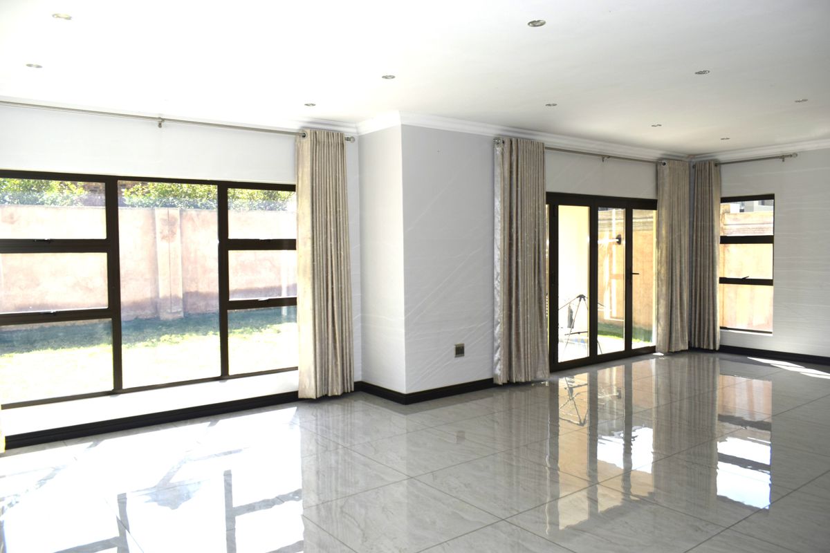 A bright and spacious open-plan living area featuring polished tiled flooring, recessed downlights, and large aluminium-framed windows and sliding doors leading to the garden.