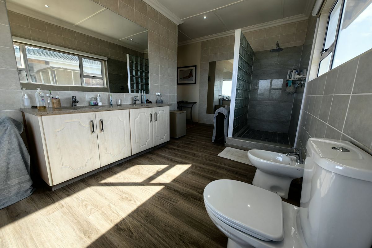 Spacious bathroom featuring a double vanity, walk-in shower with glass block partition, bidet, and toilet, finished with neutral wall tiles and wood-look flooring.