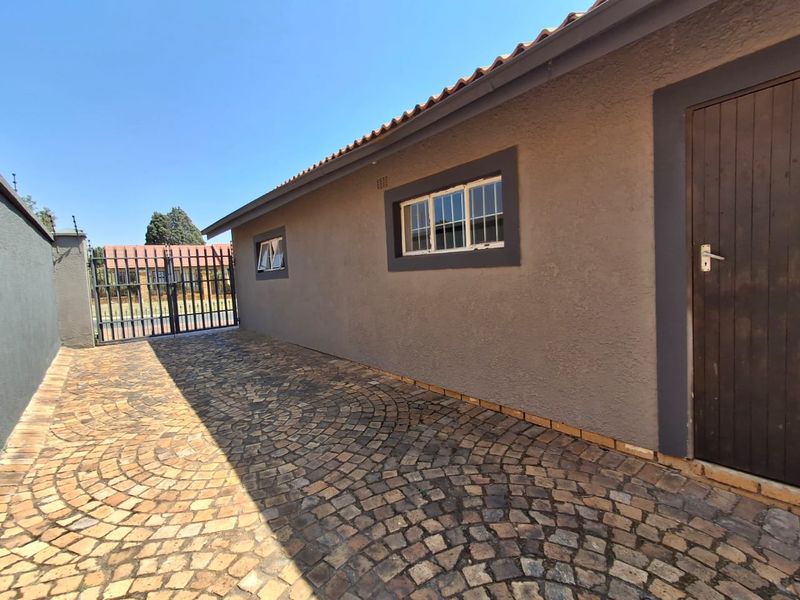 A side view of the property featuring durable cobblestone paving, a secure metal gate, and well-maintained plastered exterior walls with security-fitted windows.
