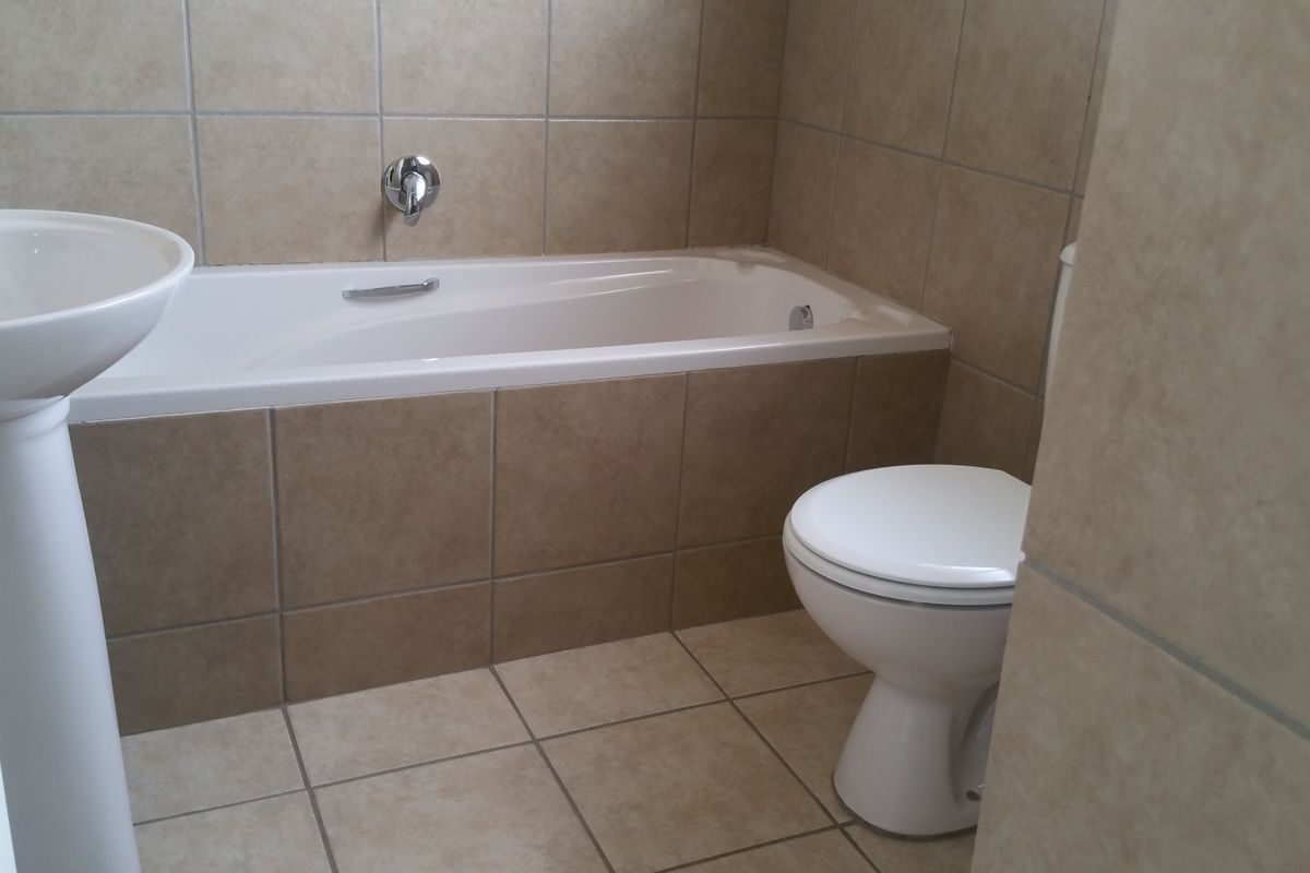 A functional bathroom featuring a pedestal basin, a built-in bathtub with wall-mounted mixer, and a standard toilet, finished with neutral ceramic wall and floor tiles.
