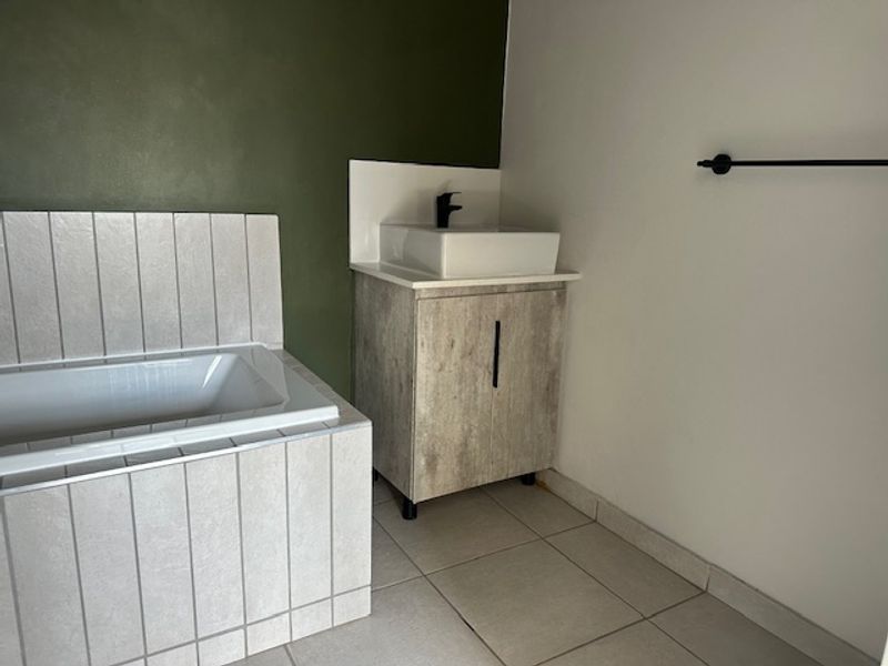 A modern bathroom featuring a tiled built-in bathtub, a wood-look vanity cabinet with a vessel sink, and a contrasting olive green accent wall.