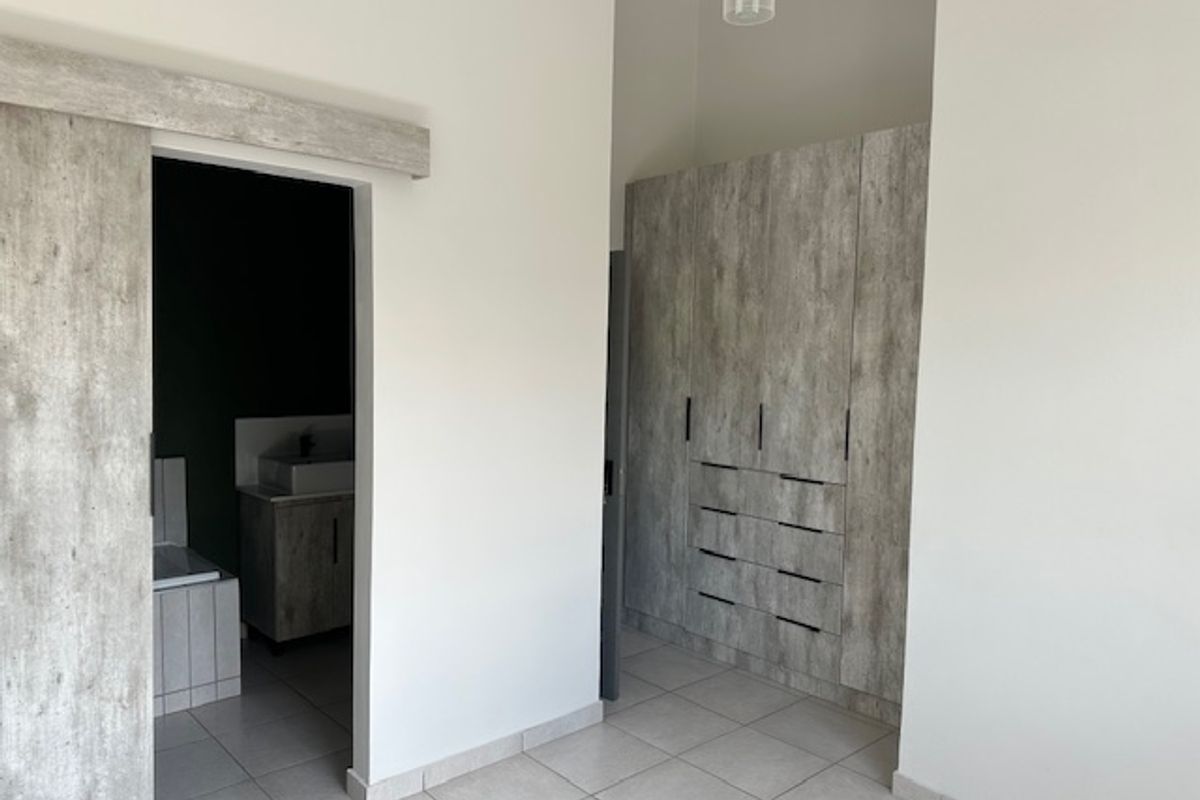 A clean, modern bedroom featuring built-in cupboards with a wood-look finish, tiled flooring, and direct access to an en-suite bathroom.