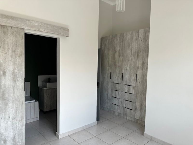 A clean, modern bedroom featuring built-in cupboards with a wood-look finish, tiled flooring, and direct access to an en-suite bathroom.