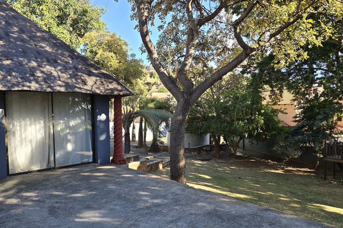 A rear view of the property featuring a thatched roof structure, concrete patio area, and a spacious garden with mature trees.
